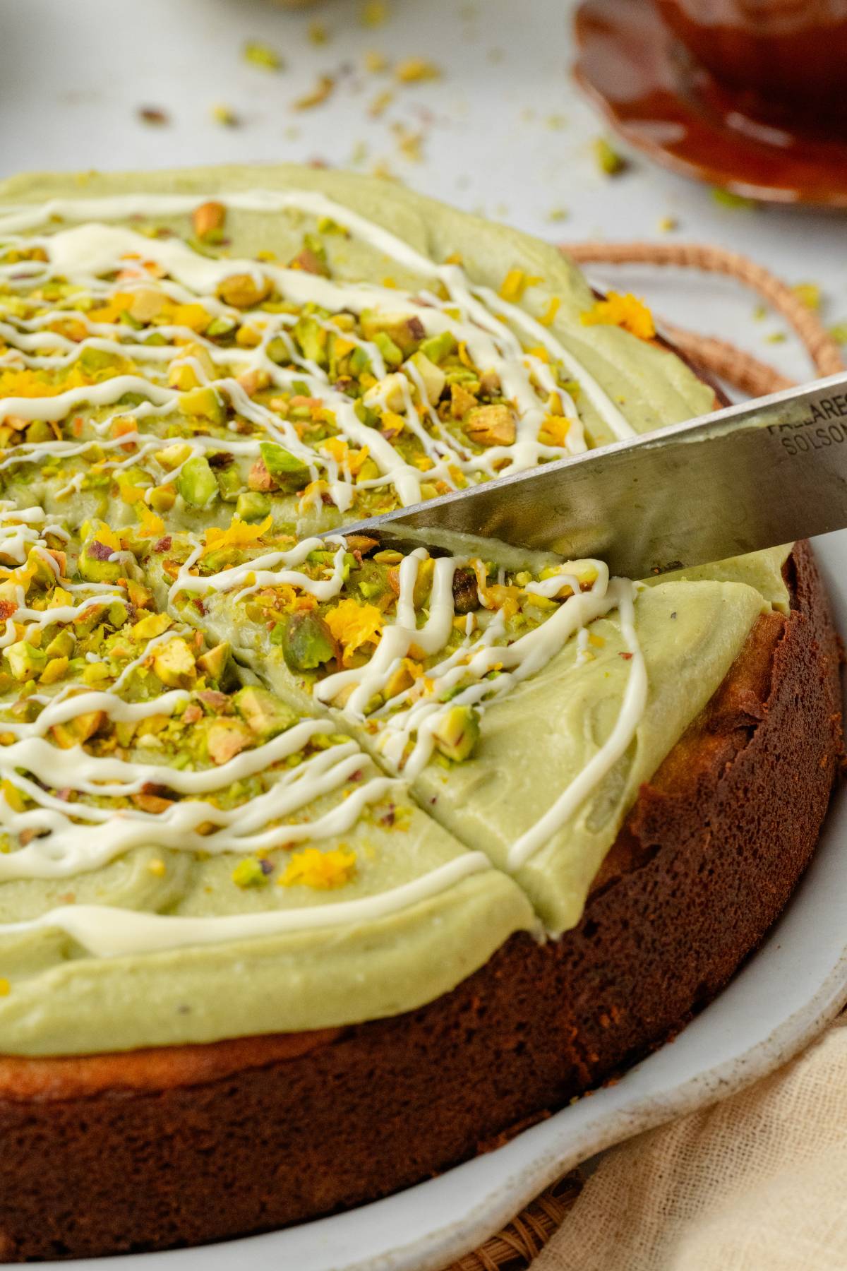 A knife slices into a pistachio ricotta cake topped with green frosting, white drizzle, and chopped pistachios.