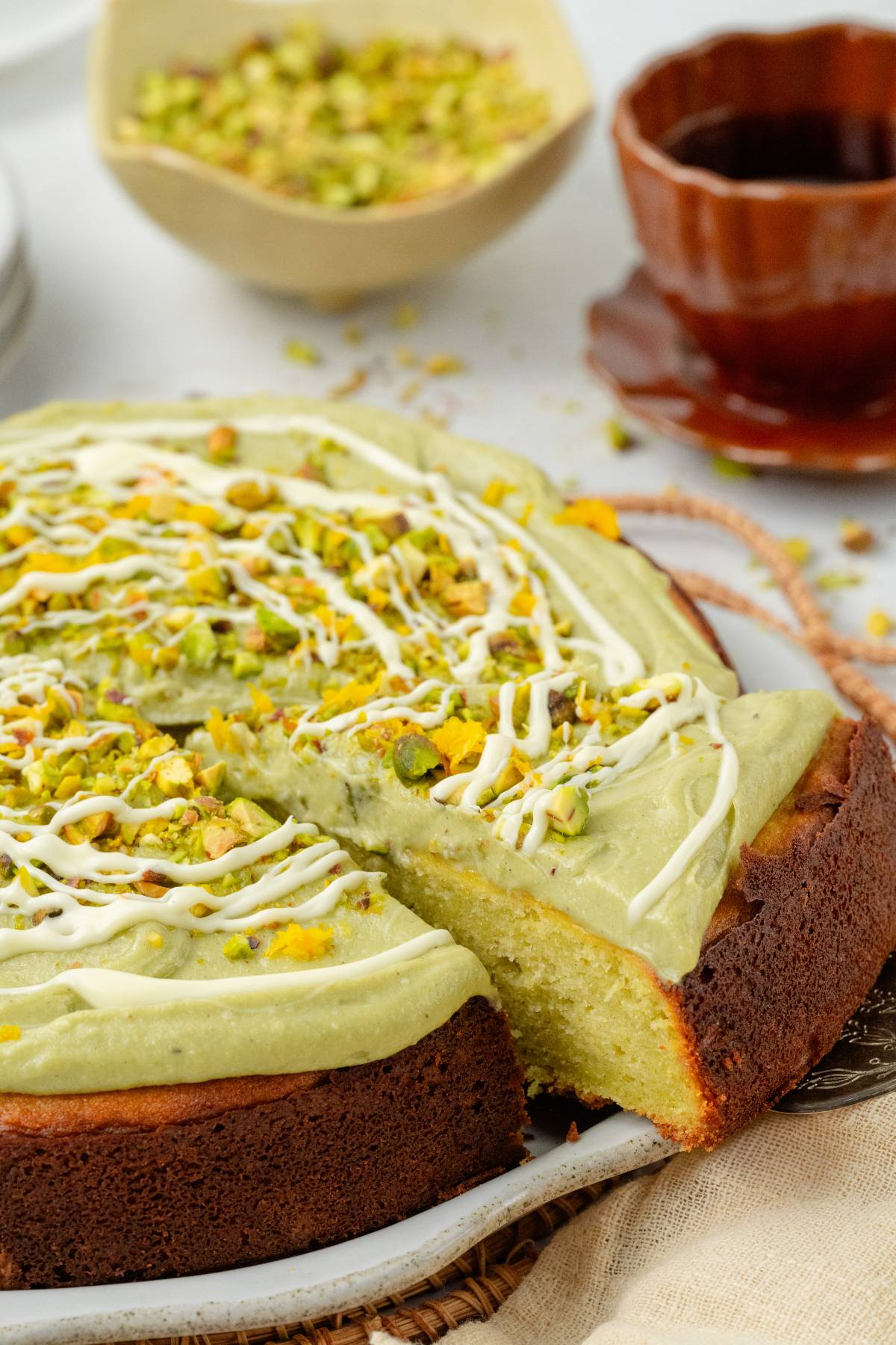 Pistachio ricotta cake with green frosting, white drizzle, and chopped pistachios, with a slice being served.