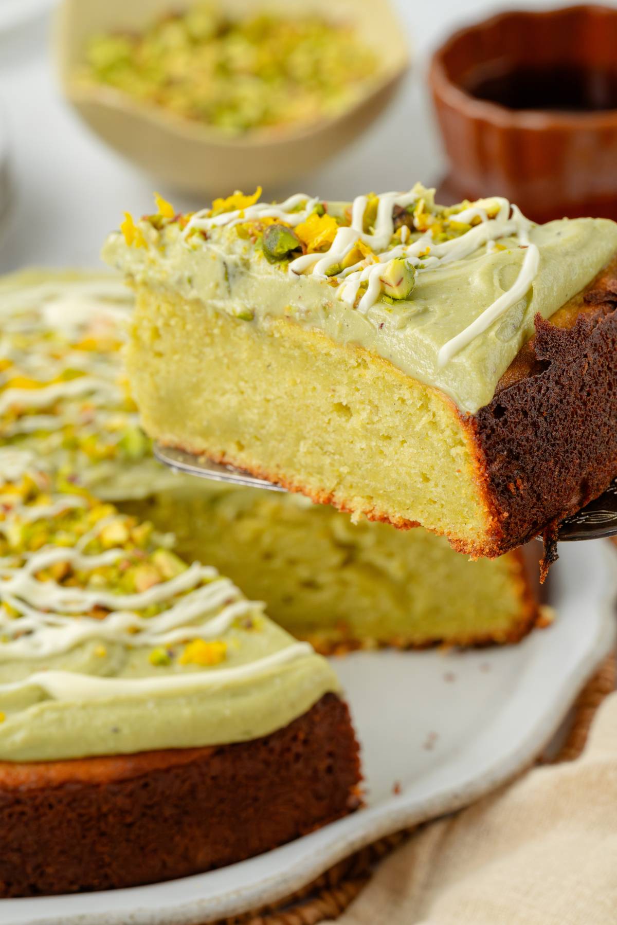 A slice of pistachio ricotta cake with green frosting and chopped pistachios is being lifted from the whole cake.