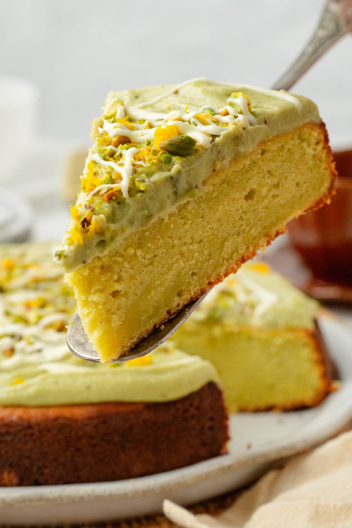A slice of pistachio ricotta cake with green frosting is lifted from a whole cake on a plate.