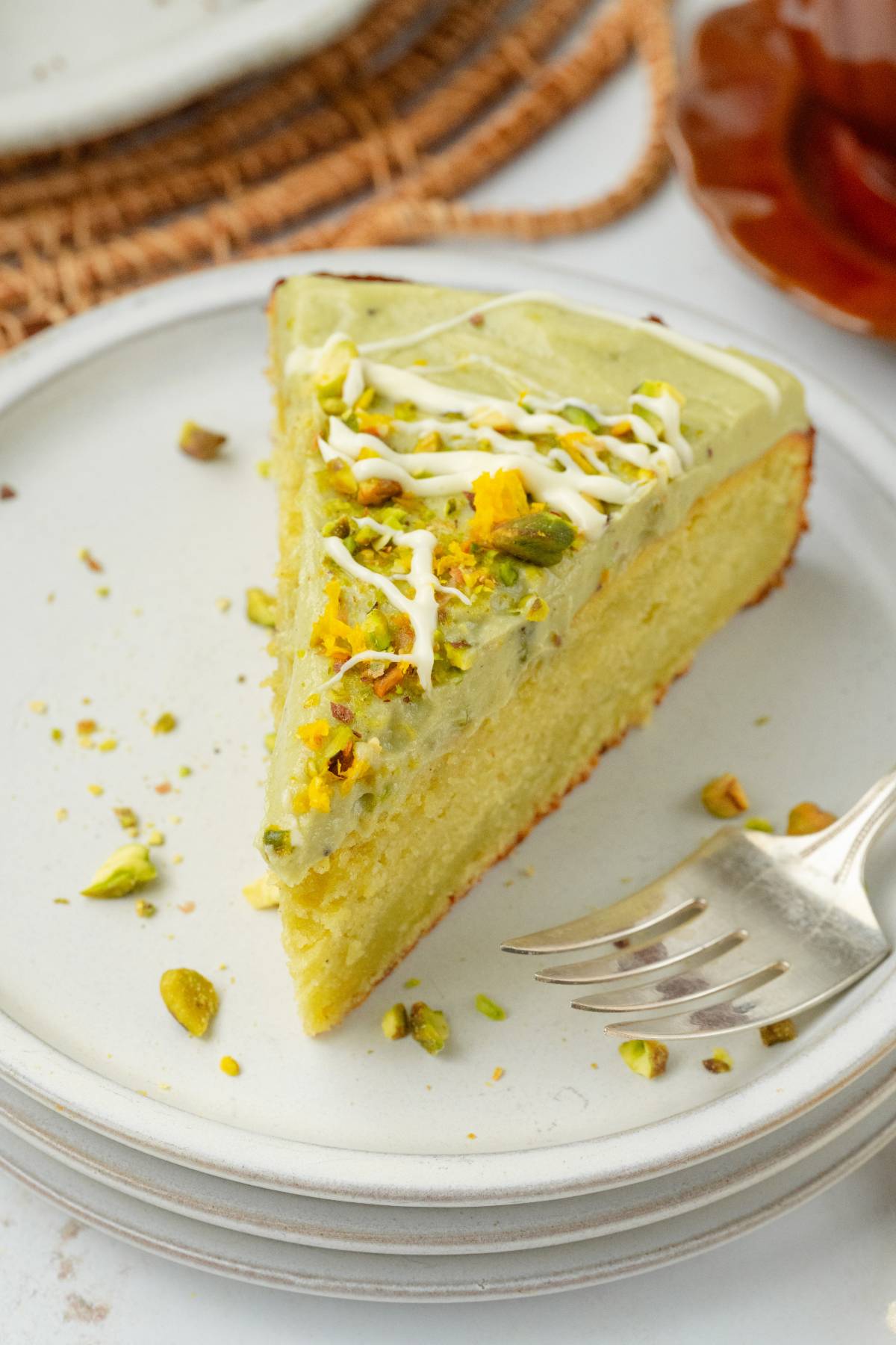 A slice of pistachio ricotta cake with green frosting and white drizzle sits on a plate, a fork placed neatly beside it.