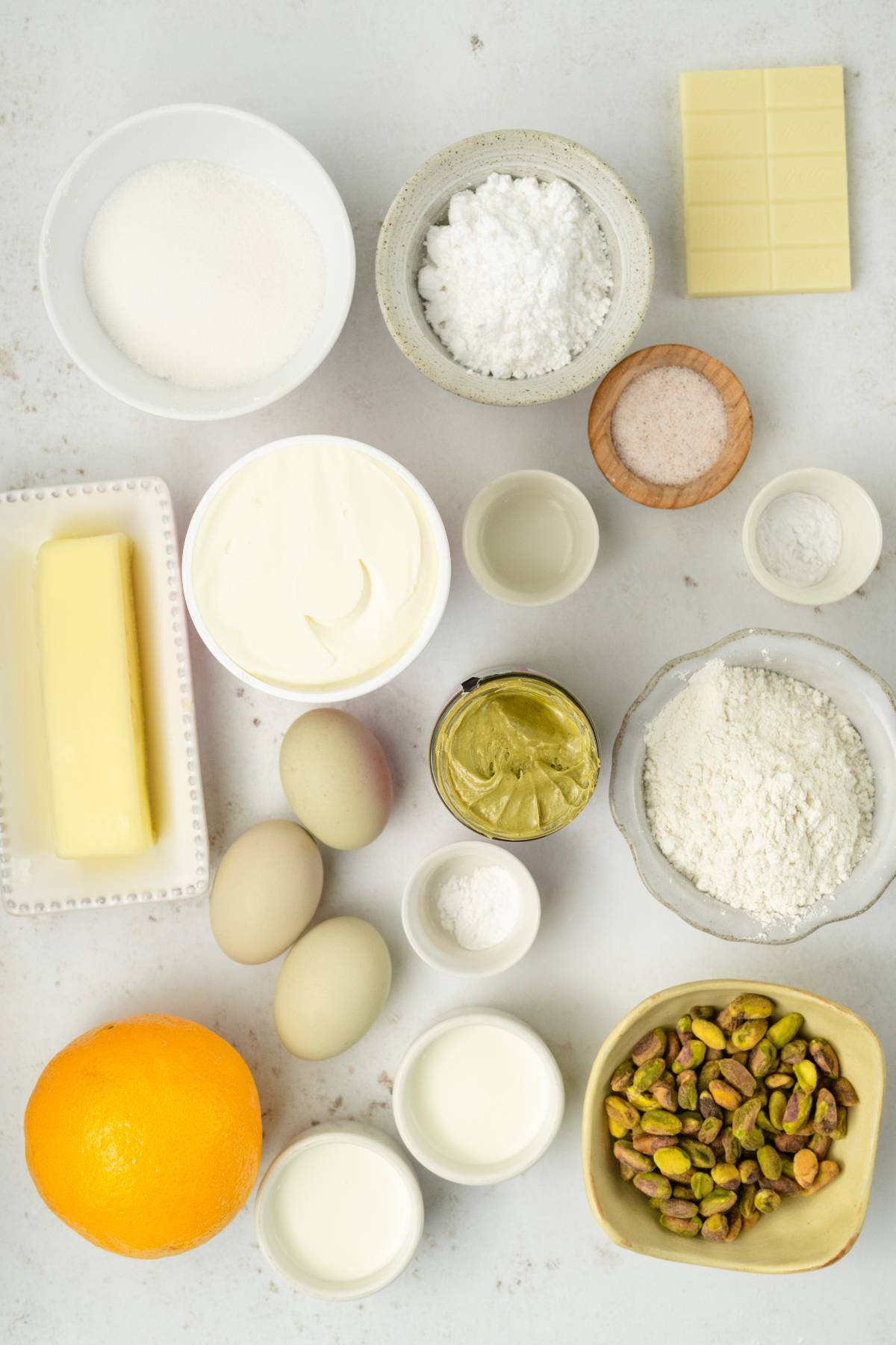 Various baking ingredients, including eggs, butter, sugar, flour, white chocolate, pistachios, and an orange, on a white surface—perfect for making a delicious pistachio ricotta cake.