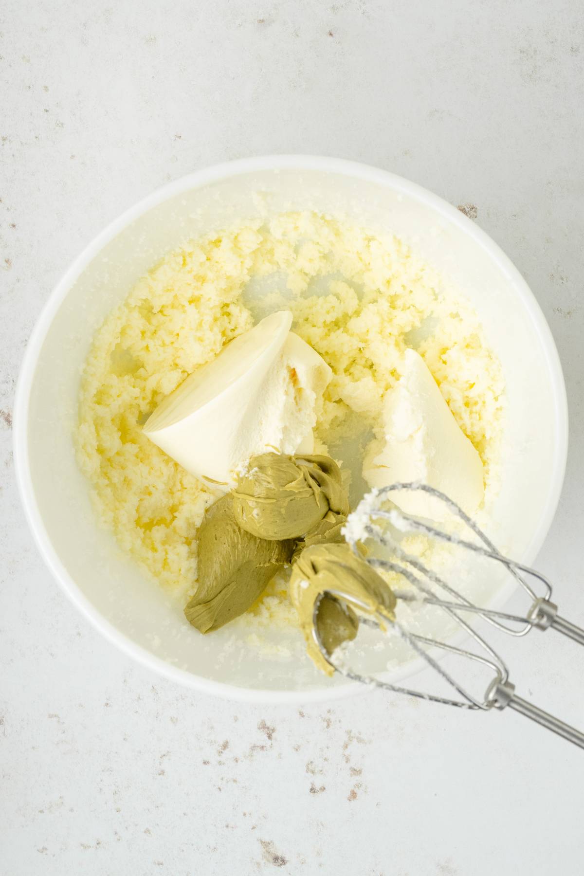 A bowl with creamed mixture, cream cheese, matcha, and flour—perfect for making a pistachio ricotta cake—sits next to a handheld electric mixer.