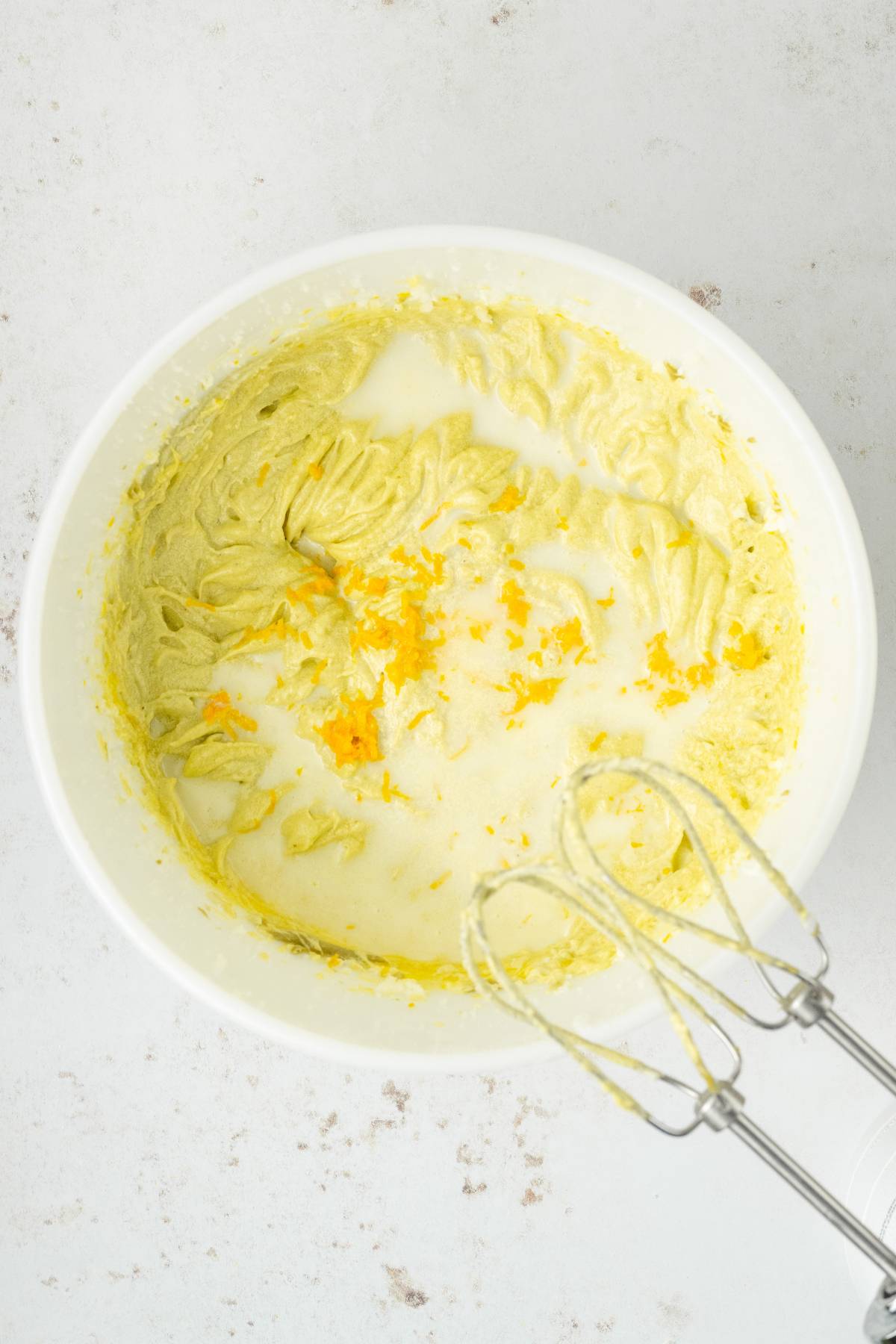 Mixing bowl with yellow batter for pistachio ricotta cake, milk, orange zest, and a hand mixer on a white surface.