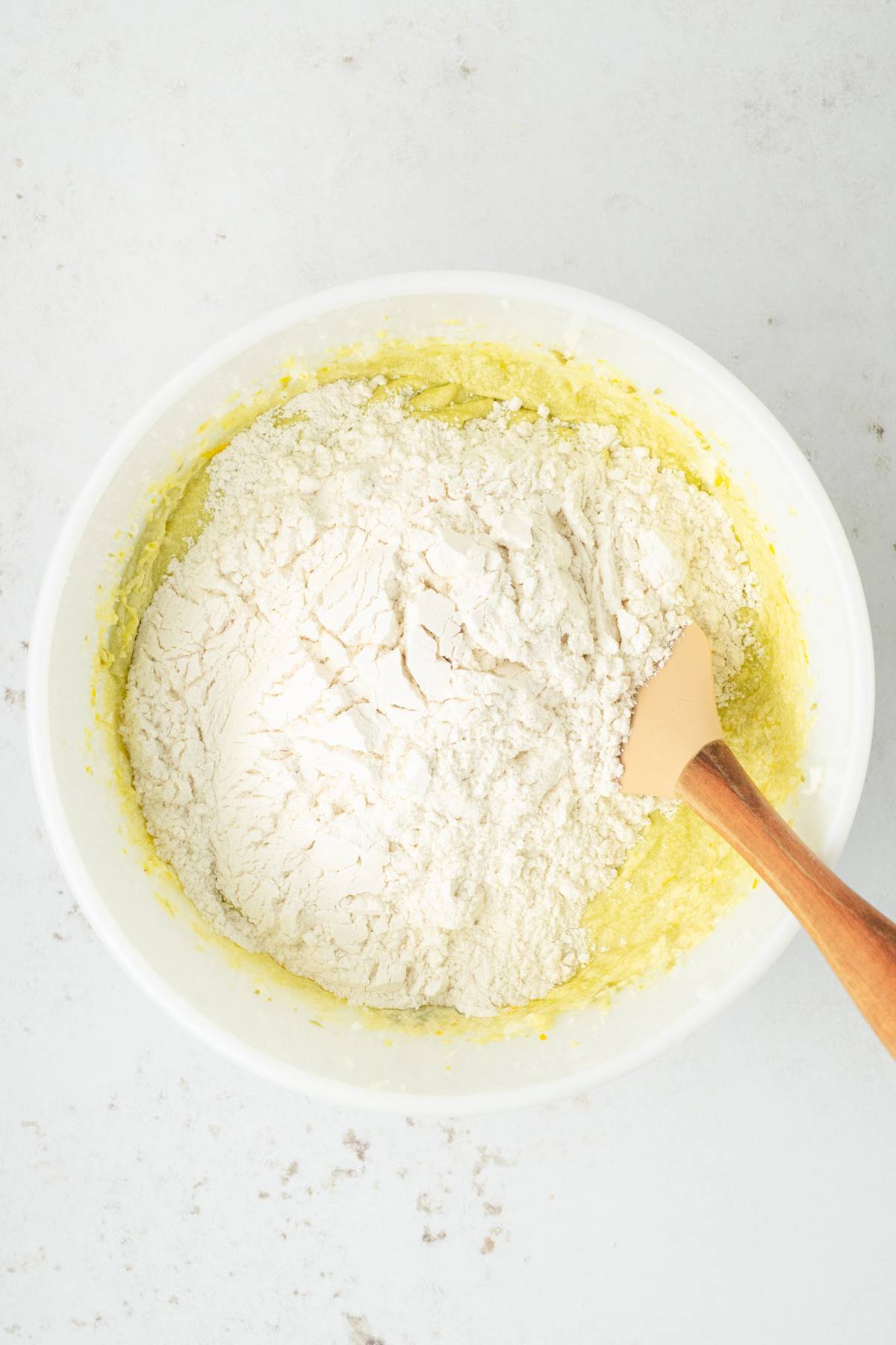 A mixing bowl with flour on top of yellow batter for a pistachio ricotta cake, with a wooden spatula resting inside.