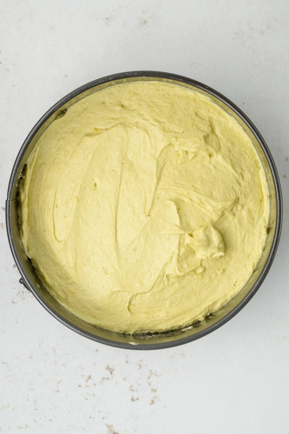 Round cake pan filled with smooth, pale yellow pistachio ricotta cake batter on a light surface.