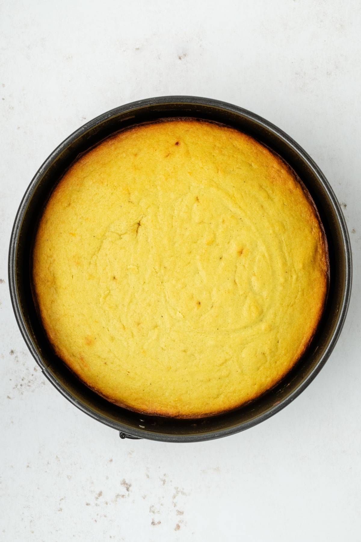 A round, golden-brown pistachio ricotta cake sits in a black baking pan on a white surface.