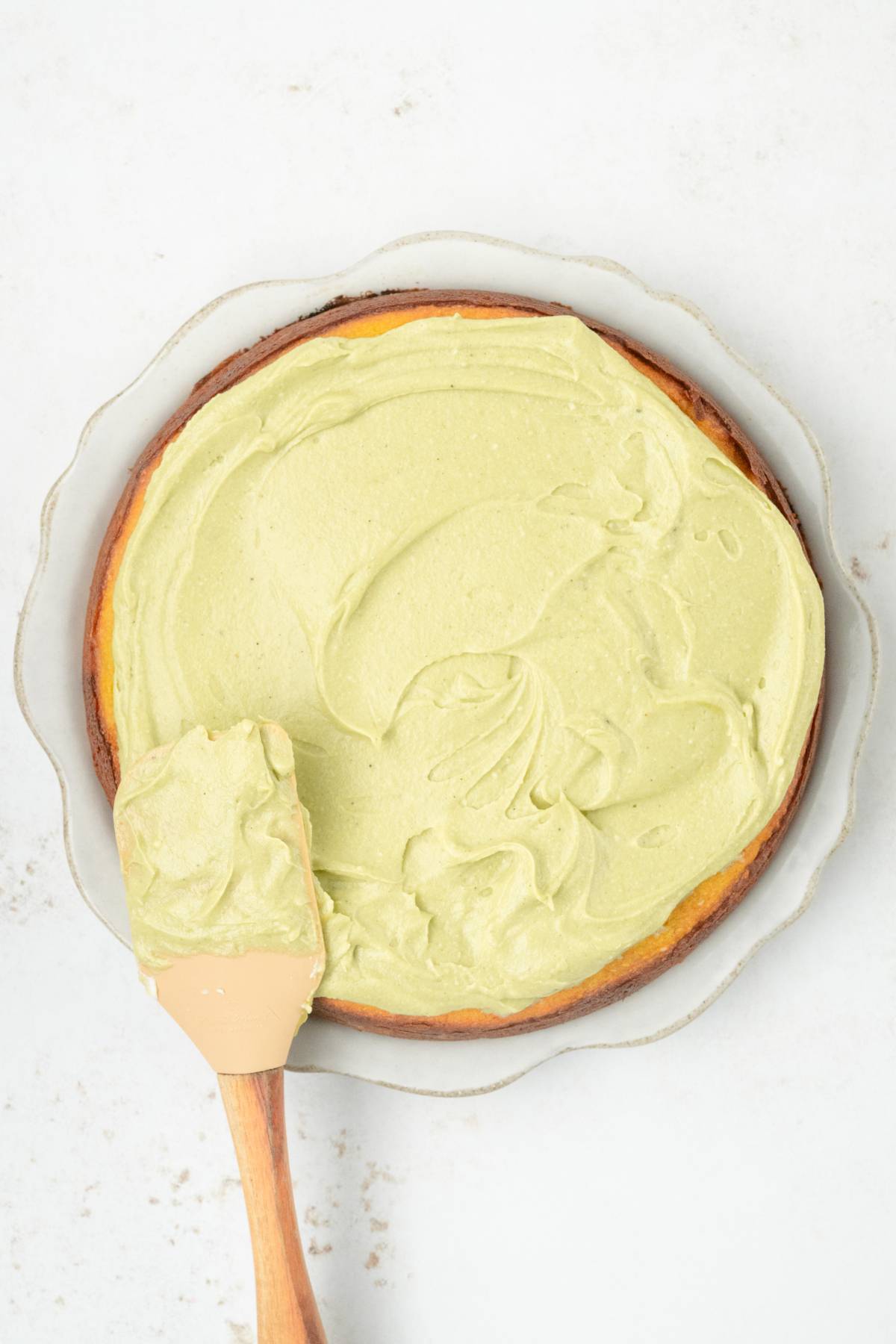 A round pistachio ricotta cake with light green frosting is being spread with a spatula on a glass plate.