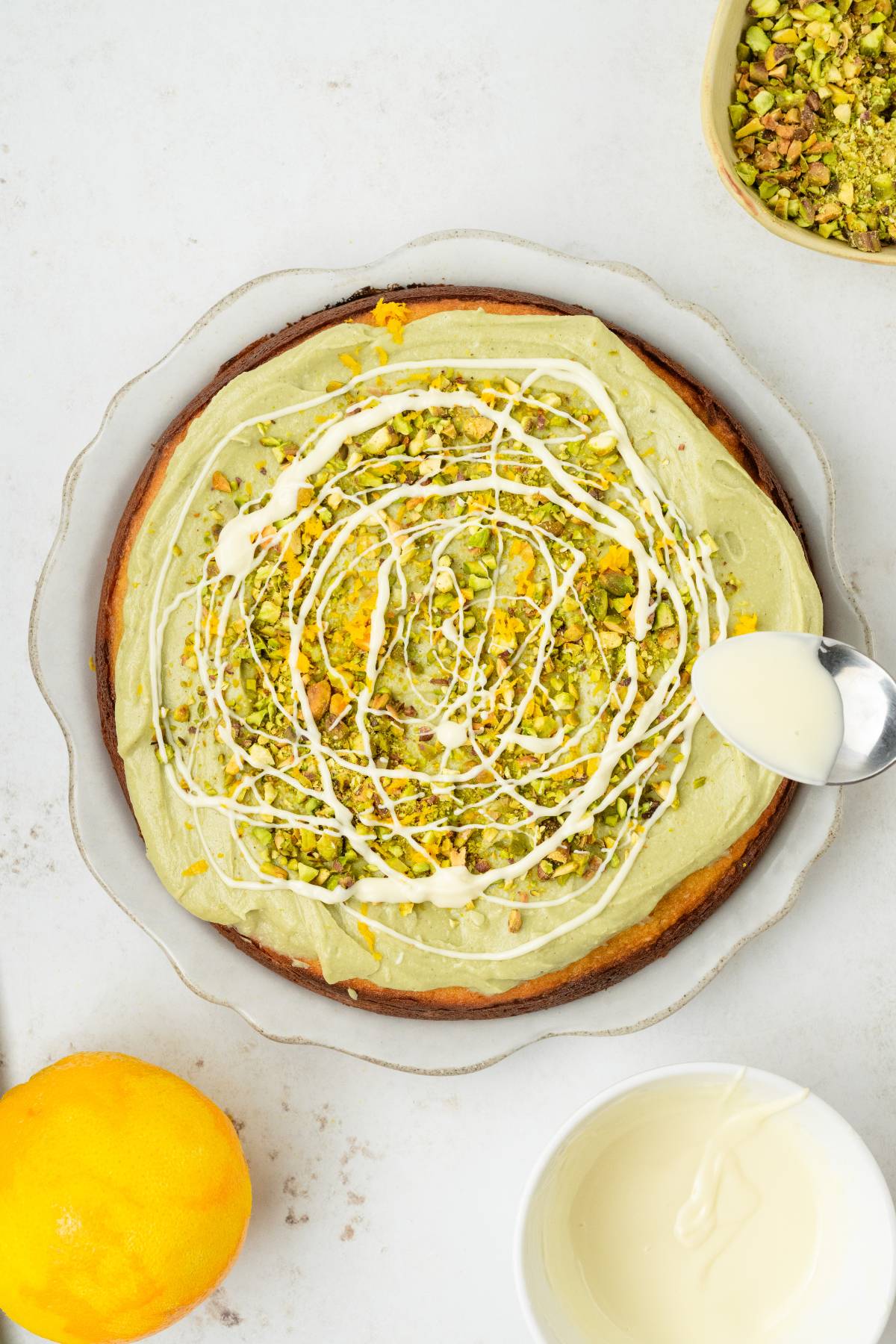 Pistachio ricotta cake topped with green frosting, chopped pistachios, and white drizzle, with a spoon adding even more drizzle.
