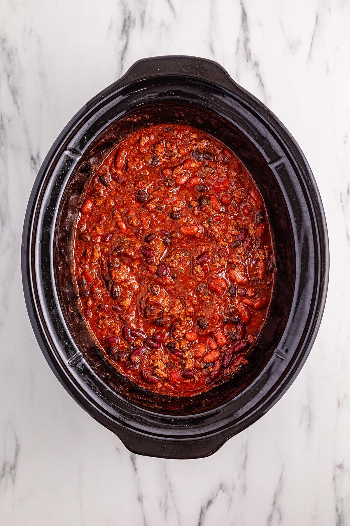 Slow cooker chili fills a pot with thick, chunky goodness, resting on a white marble countertop and viewed from above.