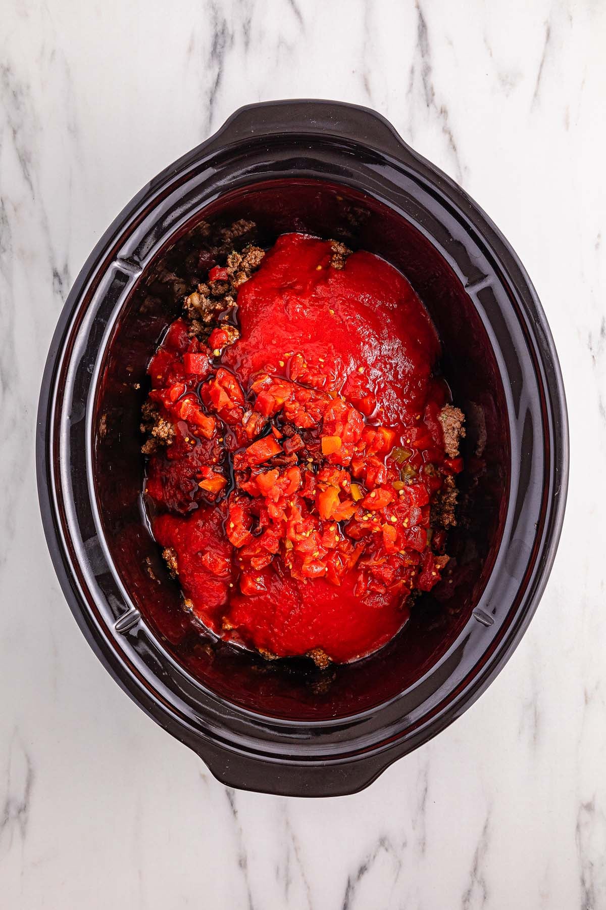 Slow cooker filled with hearty slow cooker chili, featuring ground beef, tomato sauce, and diced vegetables on a white marble surface.