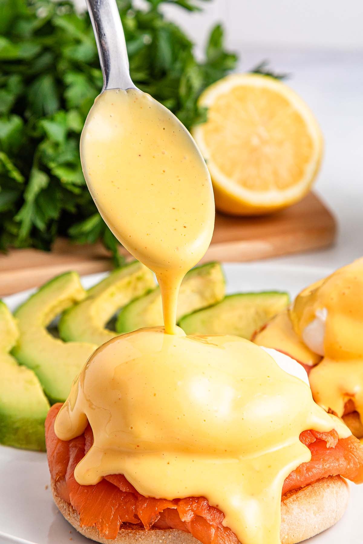 A spoon drizzles creamy hollandaise sauce over Smoked Salmon Eggs Benedict, topped with avocado slices for a delicious brunch treat.