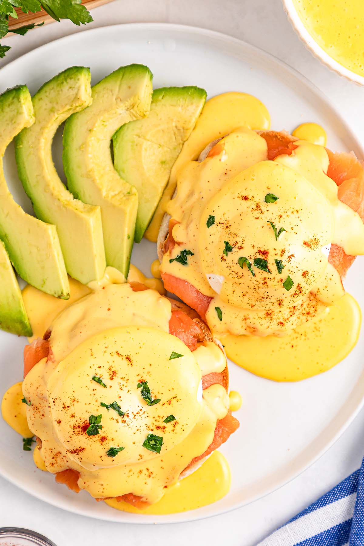 Smoked Salmon Eggs Benedict topped with hollandaise sauce, served with sliced avocado on a white plate.