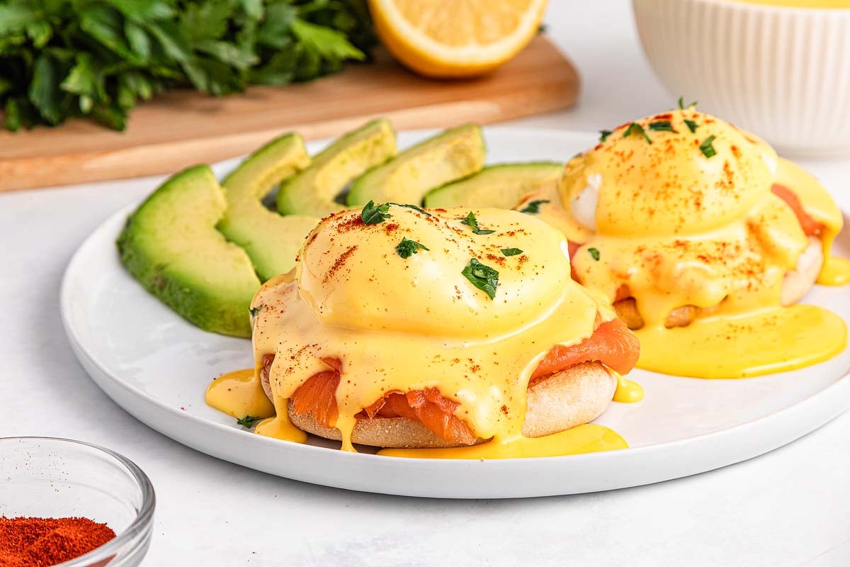 Smoked Salmon Eggs Benedict topped with creamy hollandaise sauce, served alongside fresh avocado slices on a white plate.