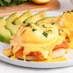 Smoked Salmon Eggs Benedict with hollandaise sauce on English muffins, served with sliced avocado on a white plate.