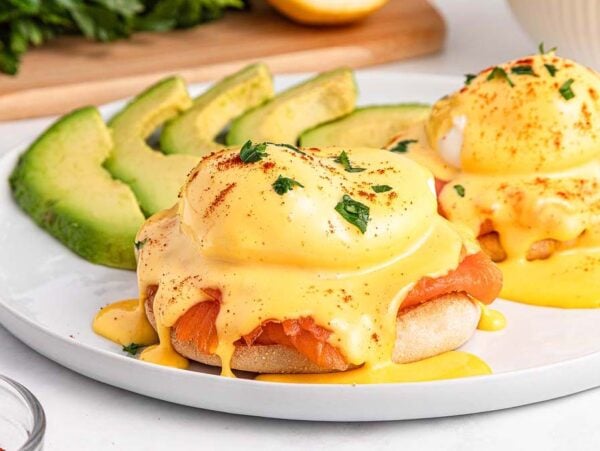 Smoked Salmon Eggs Benedict with hollandaise sauce on English muffins, served with sliced avocado on a white plate.