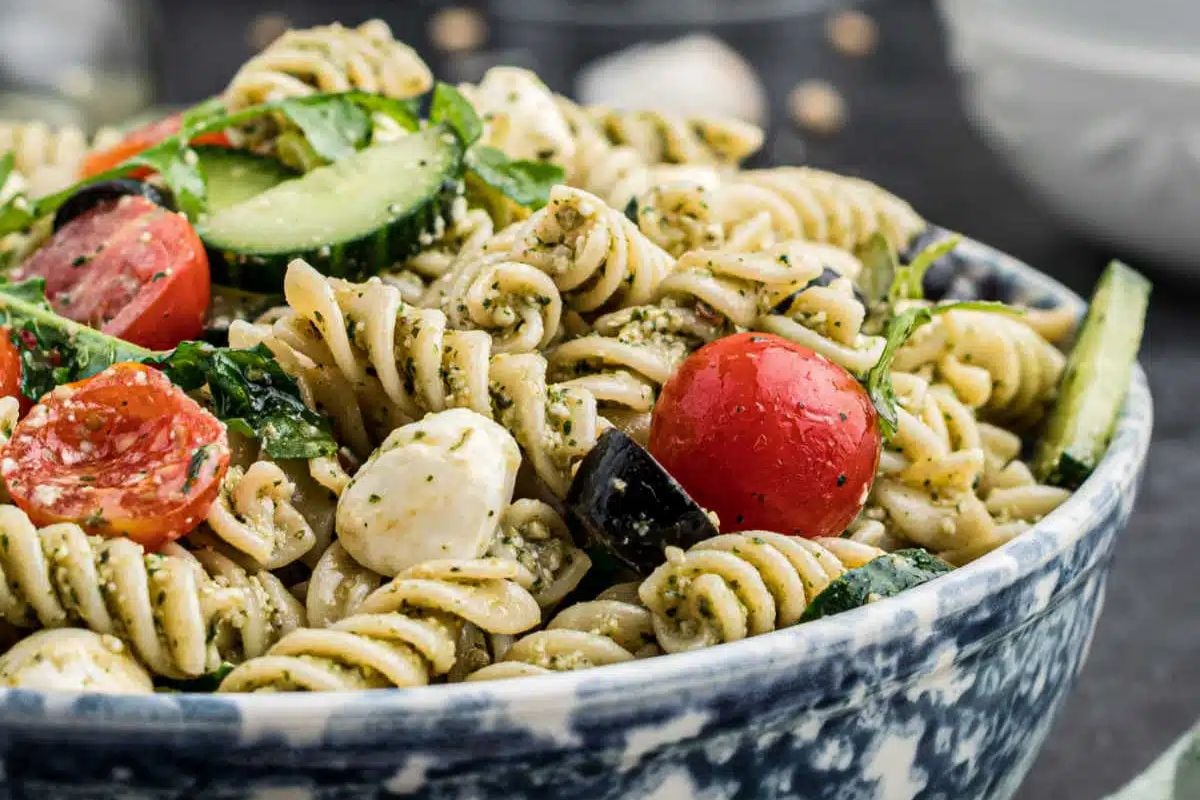 A bowl of pasta salad with rotini, cherry tomatoes, cucumber, mozzarella, olives, and leafy greens—perfect for those seeking delicious summer salad recipes.