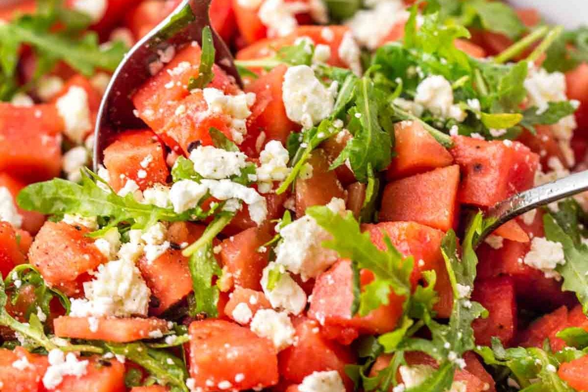 Close-up of a refreshing watermelon salad with arugula and crumbled feta cheese, perfect for summer salad recipes, served with metal utensils.