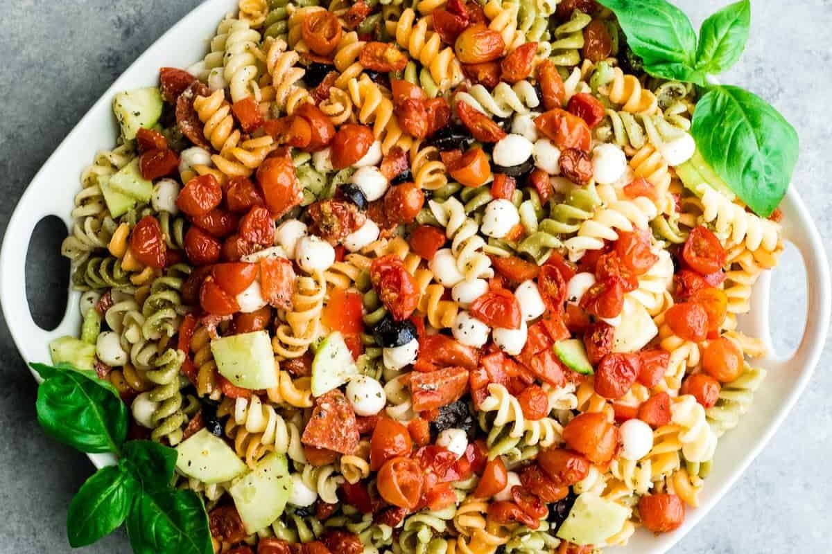 Tri-color rotini pasta salad with tomatoes, mozzarella, cucumber, olives, and fresh basil on a white platter—one of the tastiest summer salad recipes for any gathering.