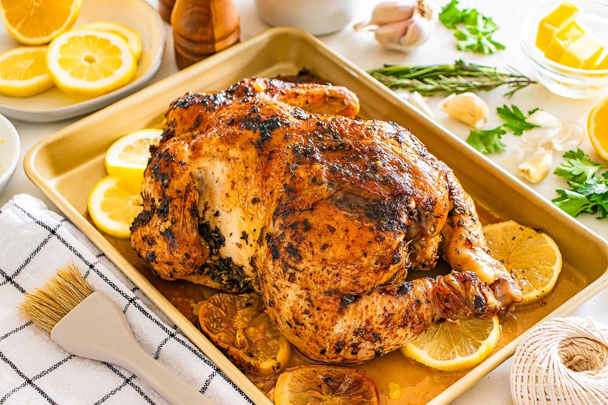 Lemon herb roasted chicken on a baking tray with lemon slices, fresh herbs, and seasonings sits on a bright kitchen countertop.
