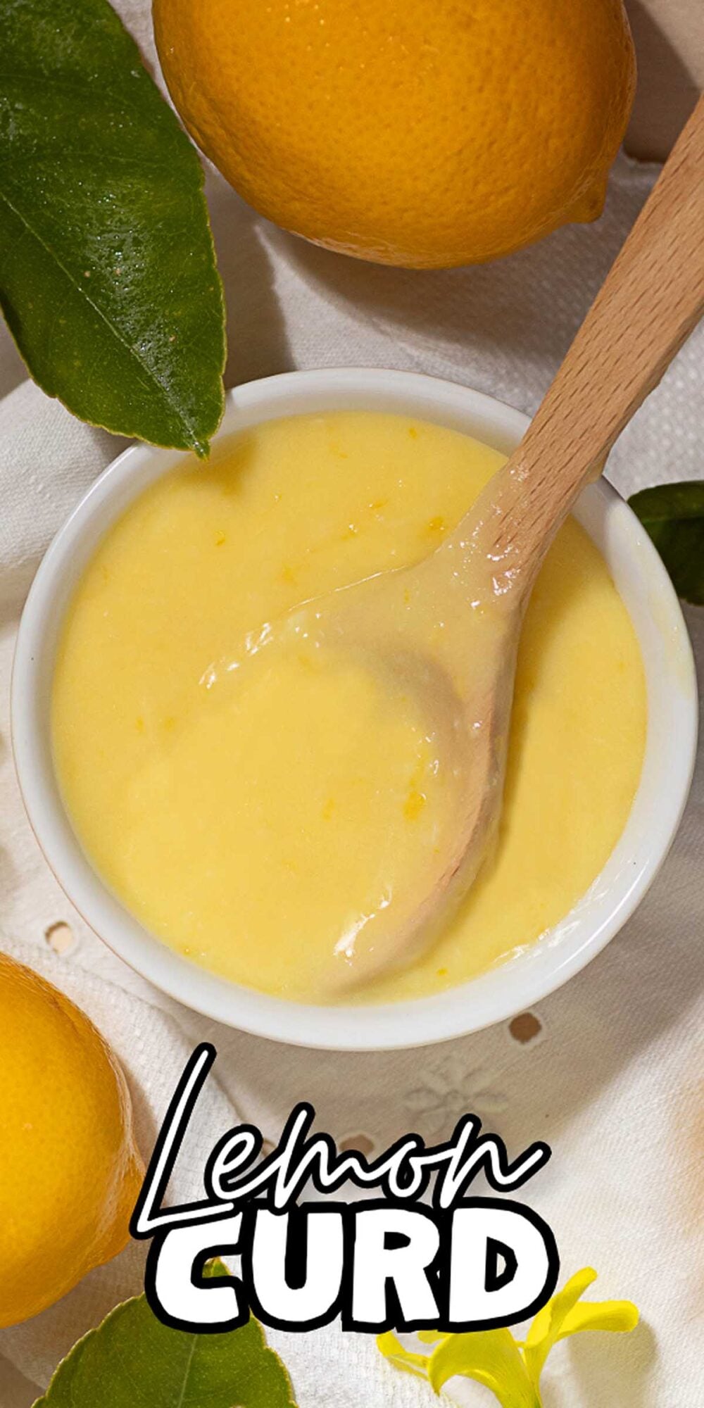 A bowl of creamy lemon curd with a wooden spoon, surrounded by fresh lemons and green leaves.
