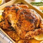 Lemon herb roasted chicken with fresh herbs and lemon slices on a baking tray, surrounded by aromatic herbs and vibrant lemon wedges.