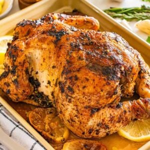 Lemon herb roasted chicken with fresh herbs and lemon slices on a baking tray, surrounded by aromatic herbs and vibrant lemon wedges.