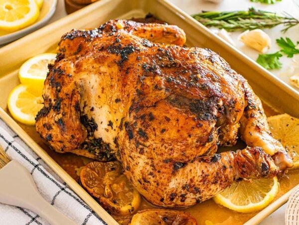 Lemon herb roasted chicken with fresh herbs and lemon slices on a baking tray, surrounded by aromatic herbs and vibrant lemon wedges.