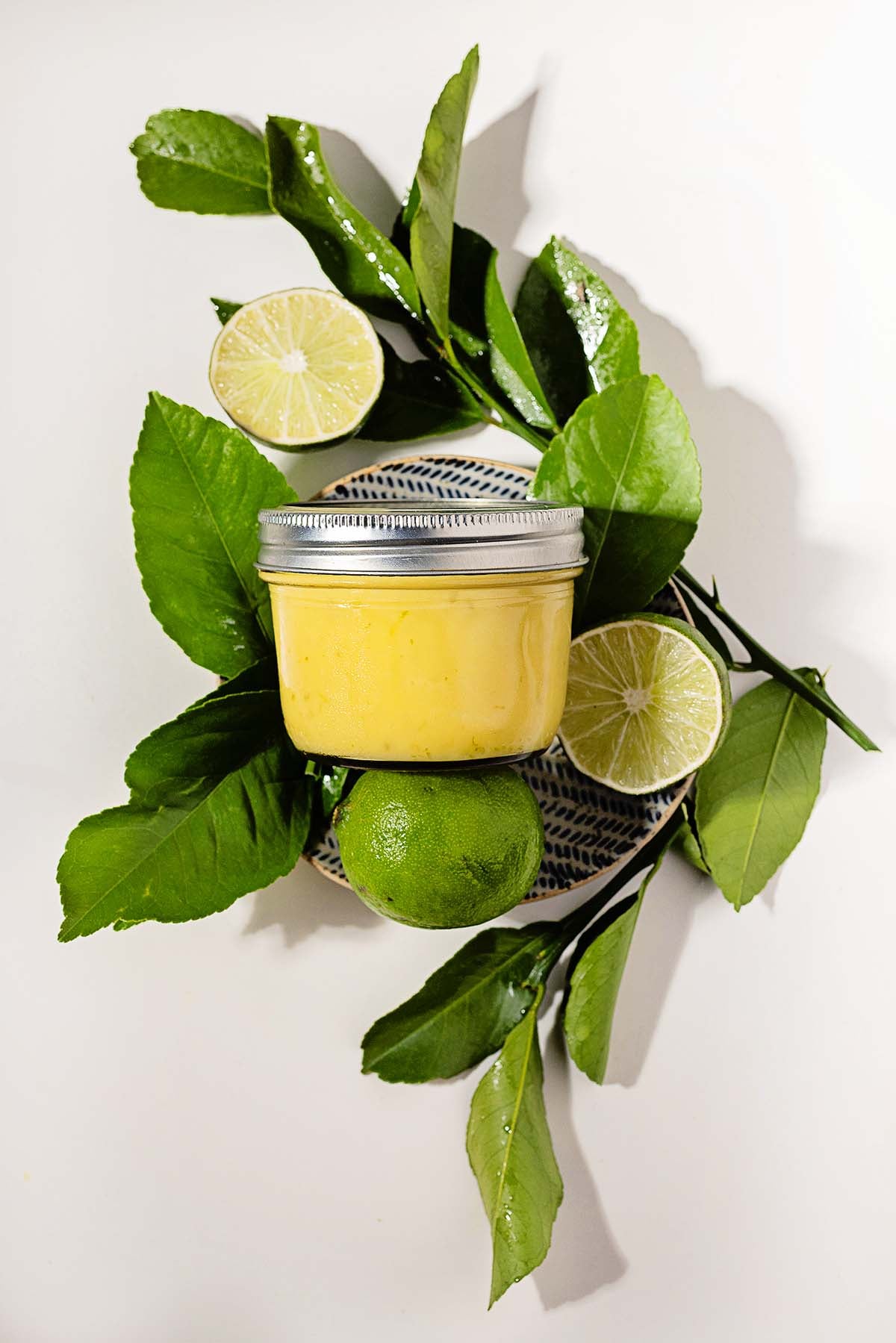 A jar of lime curd on a plate, surrounded by green leaves, a whole lime, and lime halves.