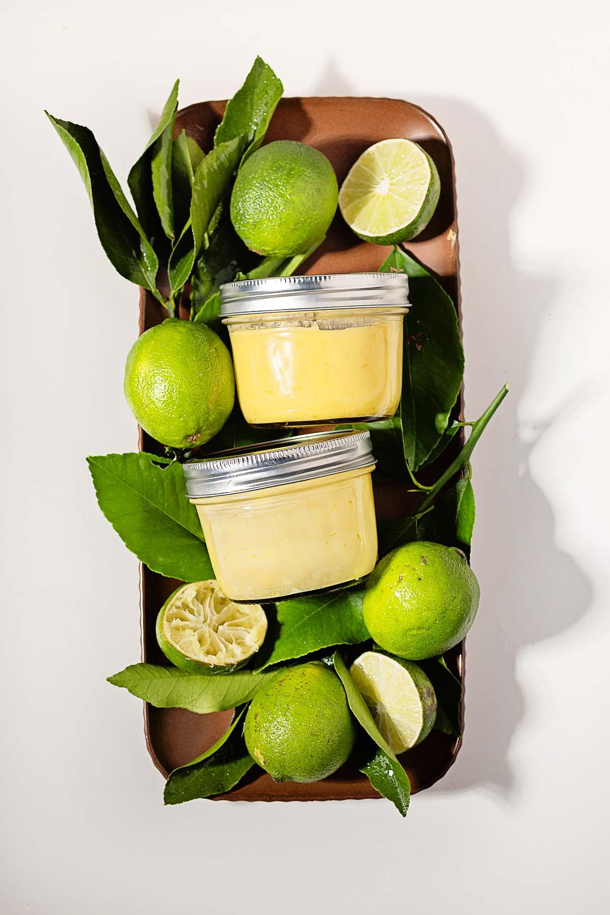 Two jars of yellow lime curd surrounded by whole and halved limes with green leaves on a brown tray.