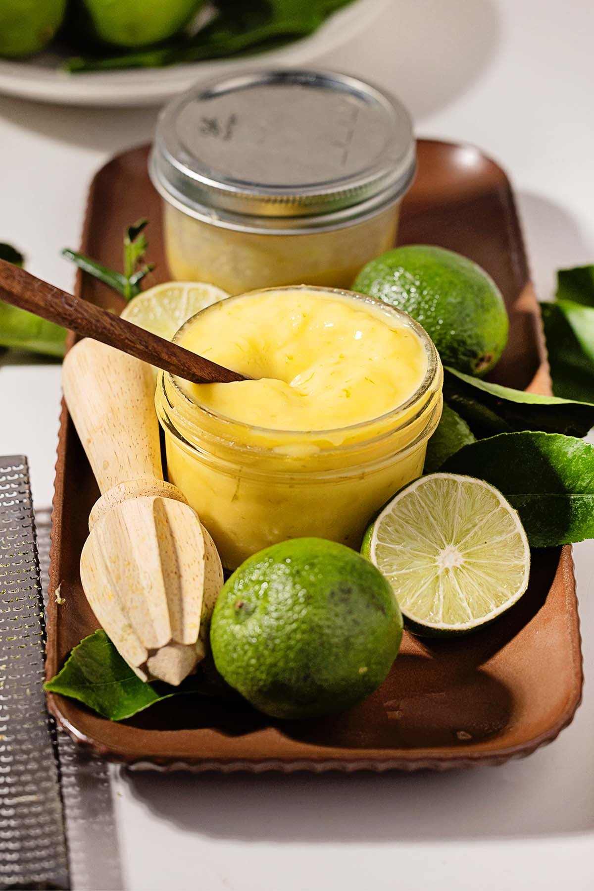 Jar of vibrant lime curd with a wooden spoon, surrounded by whole and halved limes, lime leaves, and a citrus reamer.