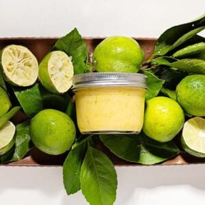 A jar of lime curd surrounded by whole and cut limes with green leaves on a tray.