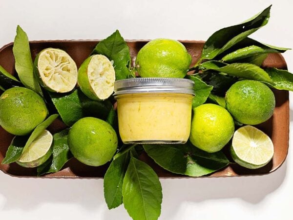 A jar of lime curd surrounded by whole and cut limes with green leaves on a tray.