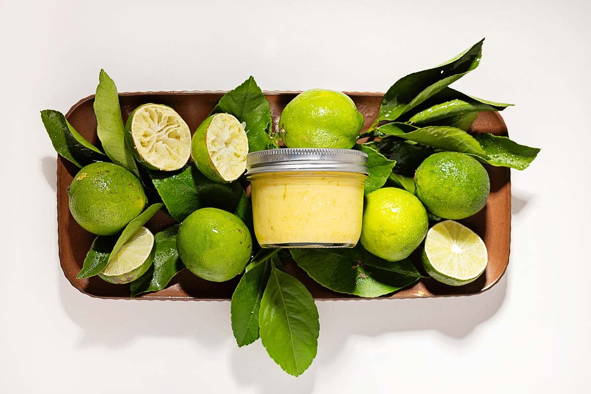 A jar of creamy lime curd spread surrounded by fresh limes, lime halves, and green leaves on a brown tray.
