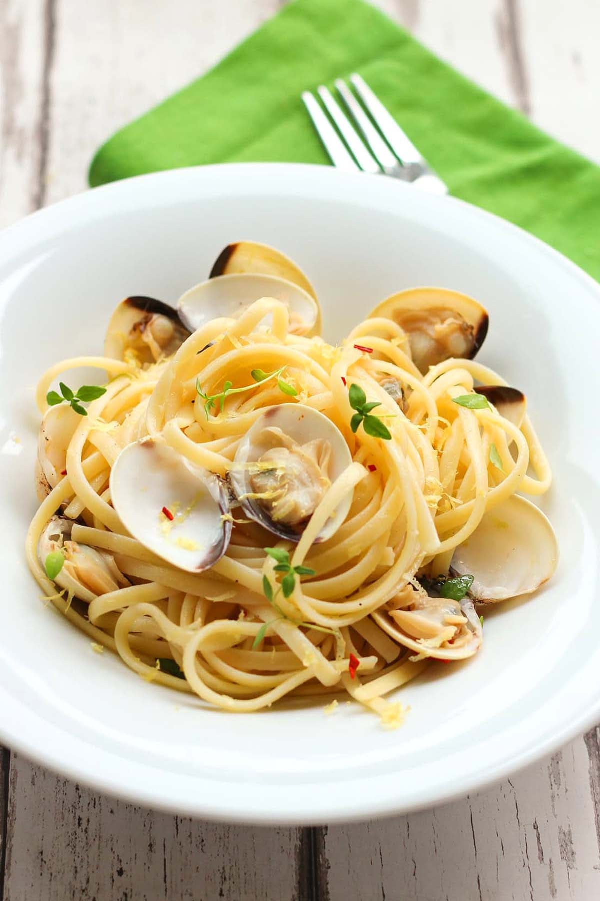 A white bowl of linguine with clams, a classic among Italian pasta recipes, garnished with herbs on a wooden table with a green napkin and fork.