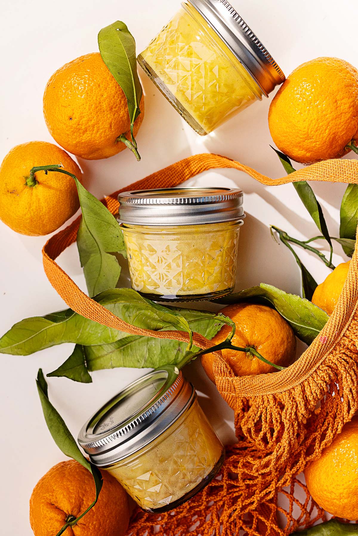 Three jars of mandarin orange curd surrounded by fresh oranges and green leaves rest on a white surface with a netted bag.