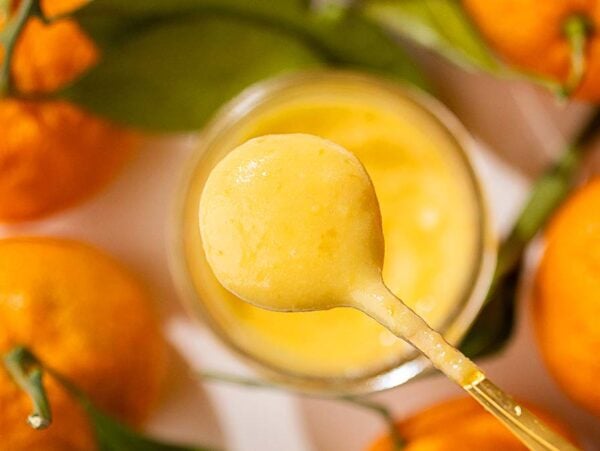 A spoon with creamy mandarin orange curd surrounded by fresh mandarins with green leaves.