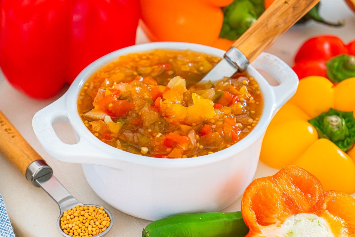 A white bowl of sweet pepper relish with a spoon, surrounded by colorful peppers and mustard seeds.