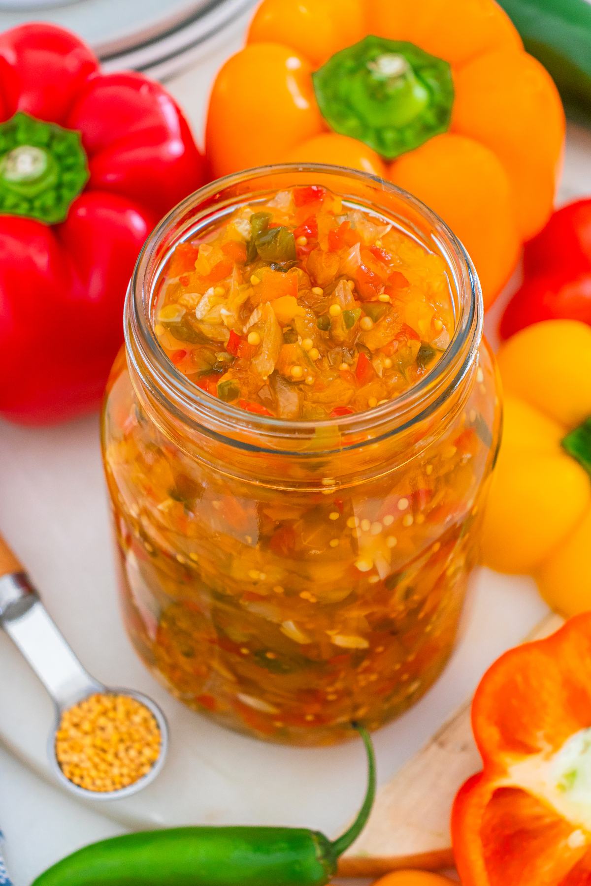 A jar of homemade relish surrounded by colorful bell peppers and a spoonful of mustard seeds.