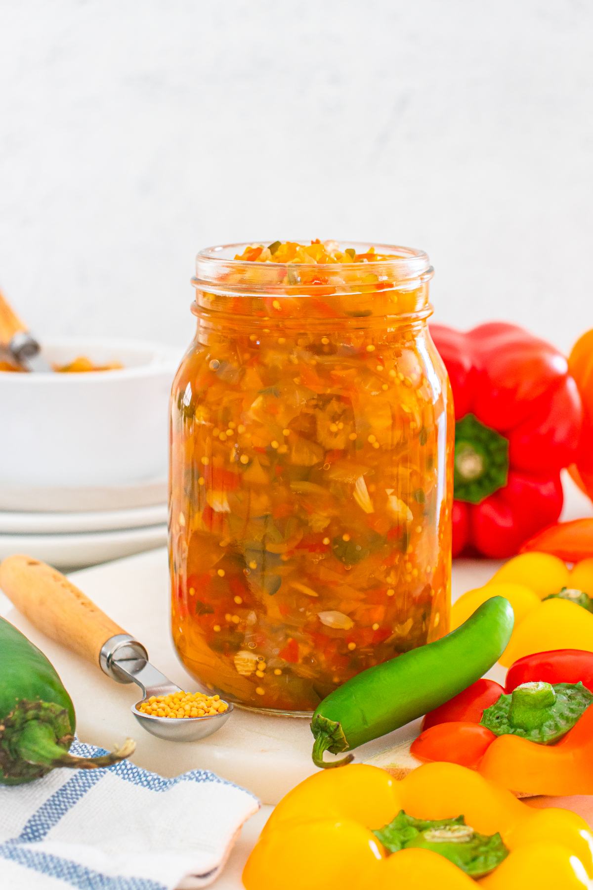 A jar of homemade relish with chopped peppers, surrounded by fresh bell peppers and a green chili.
