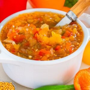 A white bowl of sweet pepper relish with a spoon, surrounded by colorful peppers and mustard seeds.