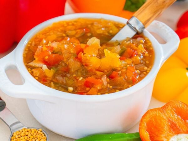A white bowl of sweet pepper relish with a spoon, surrounded by colorful peppers and mustard seeds.