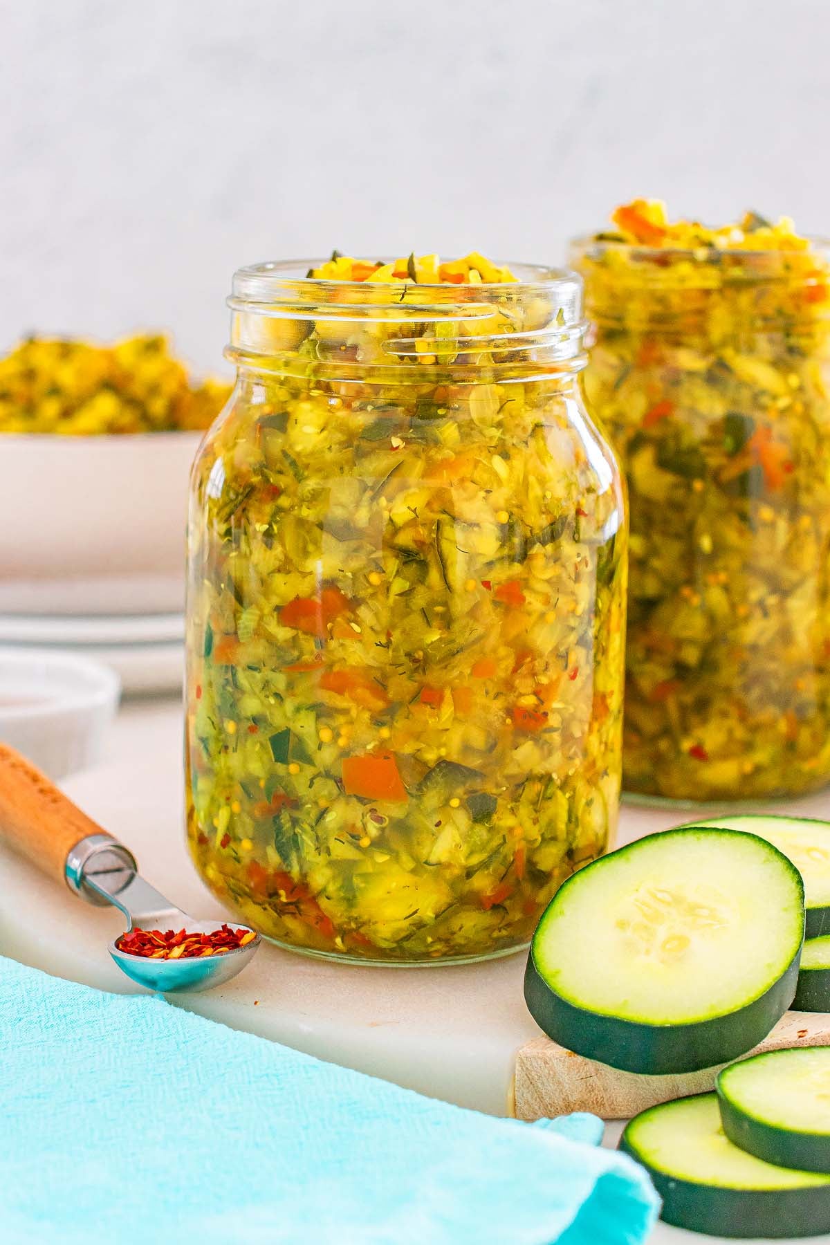 Two jars of homemade pickle relish with chopped vegetables sit beside sliced cucumbers and a spoon of red pepper flakes.