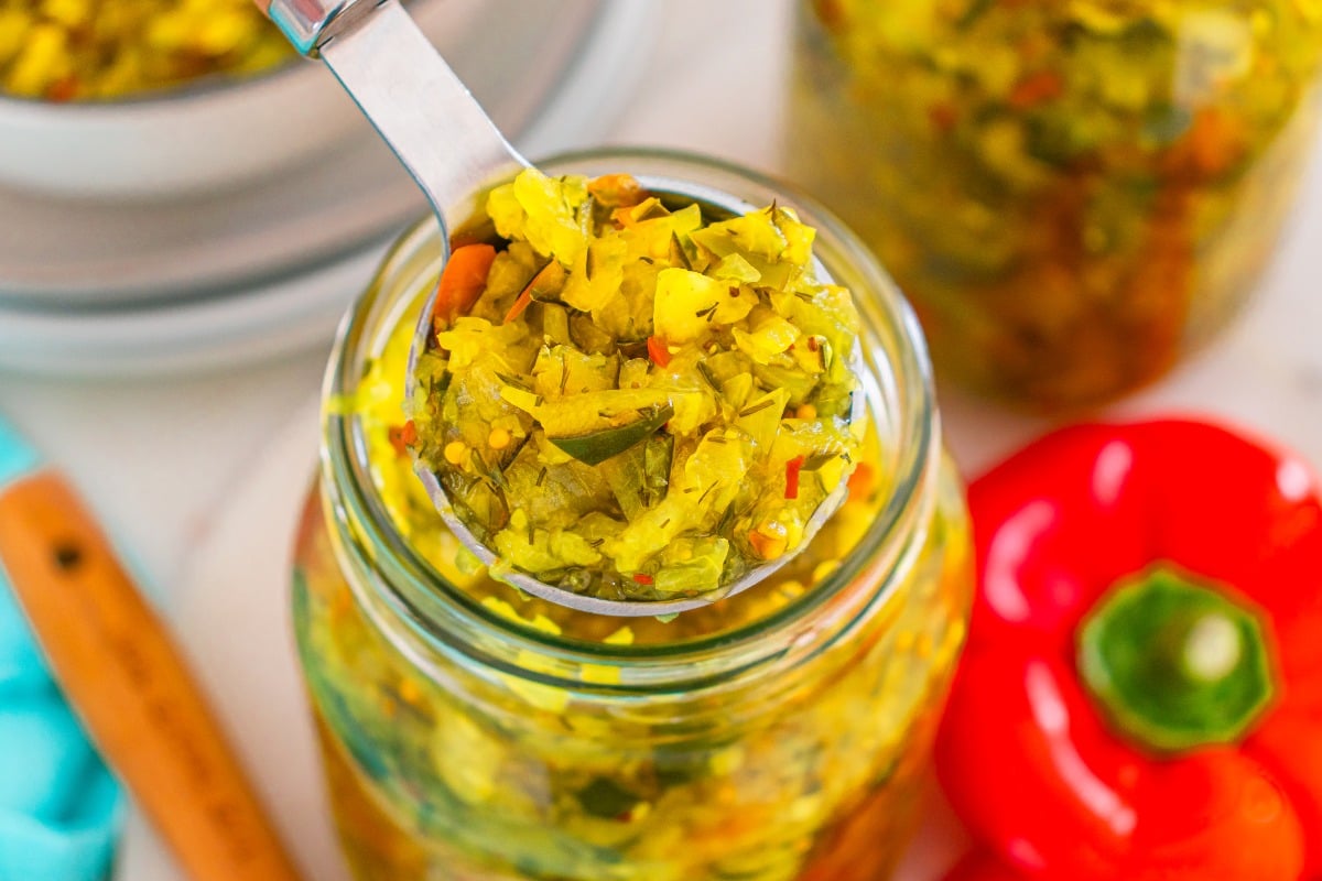 A spoon scoops yellow-green homemade relish from a glass jar, with a red bell pepper nearby.