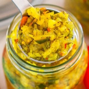 A spoonful of homemade relish being lifted from a glass jar, with a red bell pepper nearby.