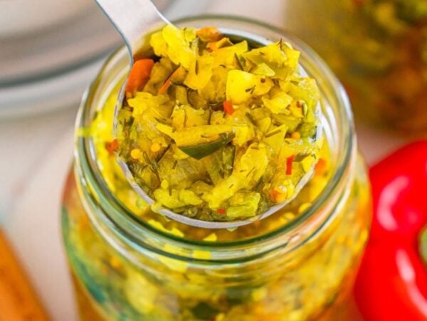 A spoonful of homemade relish being lifted from a glass jar, with a red bell pepper nearby.