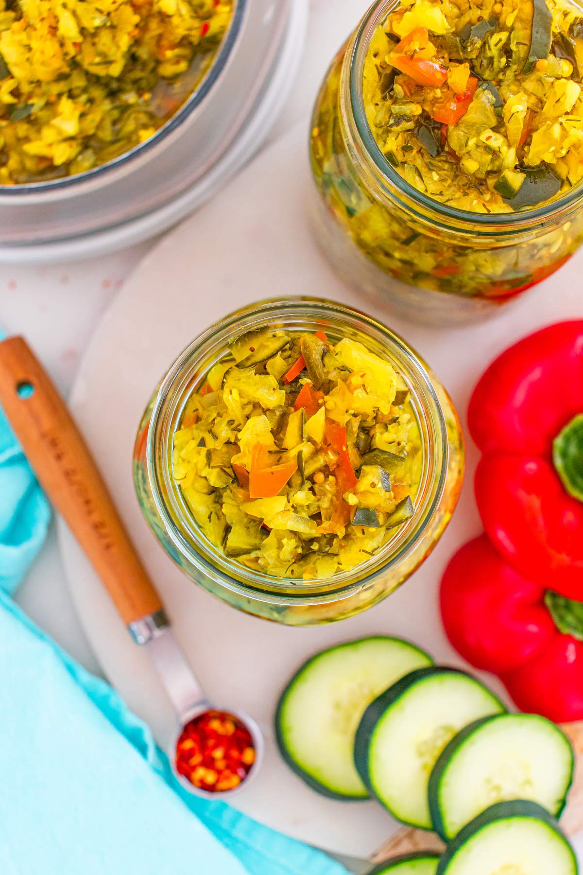 Overhead view of jars filled with homemade pickle relish, surrounded by cucumber slices and red bell pepper.