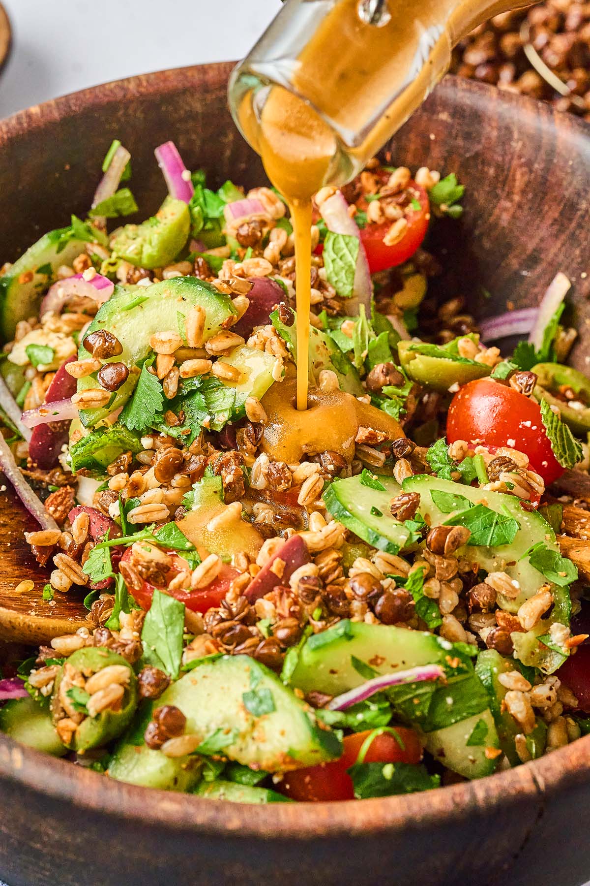 Dressing being poured onto a crispy lentil salad with grains, avocado, tomatoes, cucumbers, and herbs in a wooden bowl.