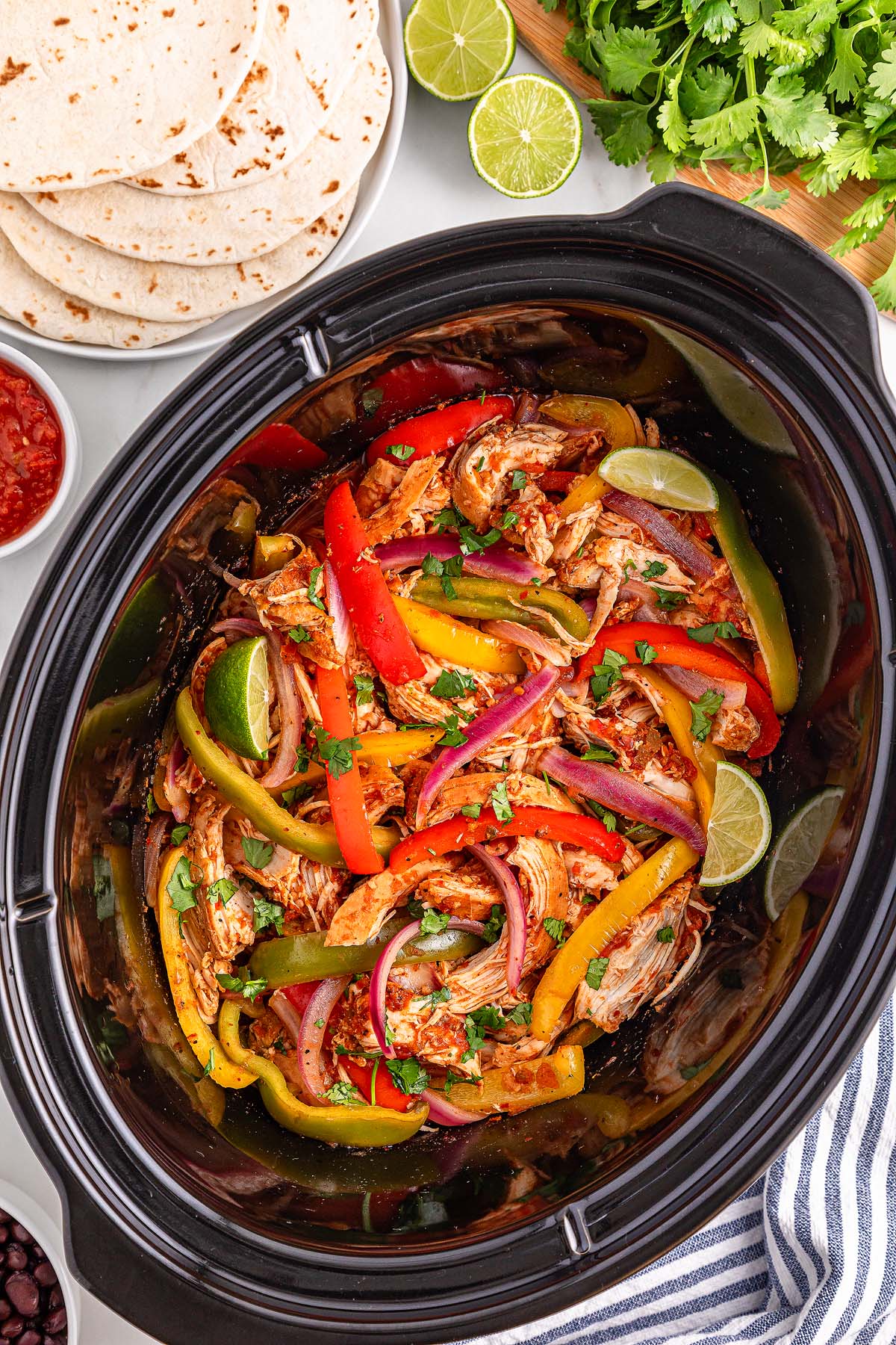Slow Cooker Chicken Fajitas featuring shredded chicken, bell peppers, and onions simmer together in a slow cooker, with warm tortillas, fresh limes, and vibrant cilantro served on the side.