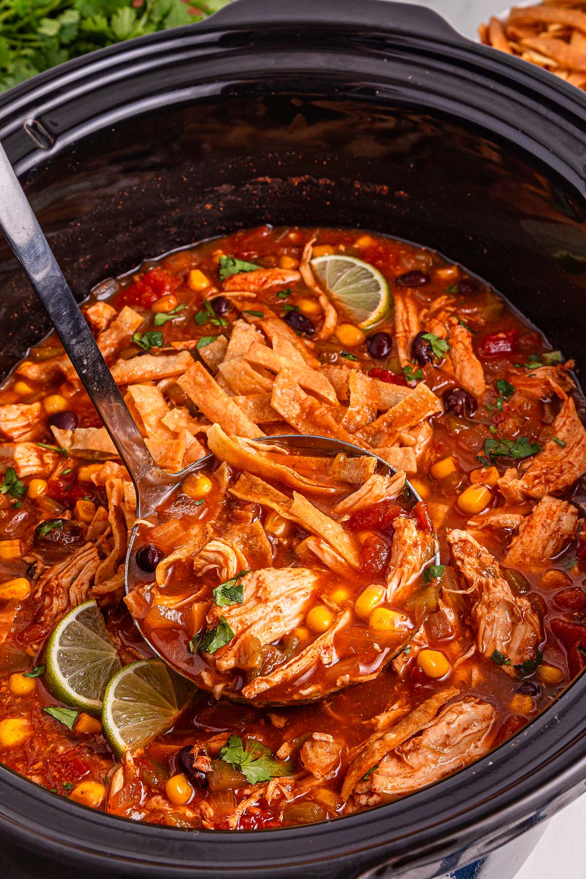A ladle scoops hearty Slow Cooker Chicken Tortilla Soup, brimming with corn, beans, lime slices, and crispy tortilla strips from a bubbling slow cooker.
